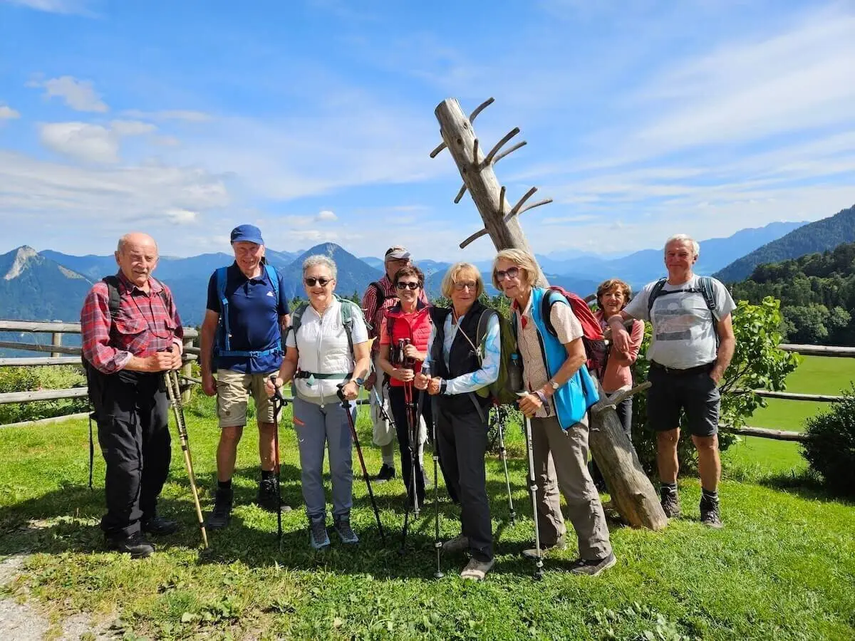 Neun Wandernde stehen auf Wiese vor Holzskulptur und Zaun, dahinter Hügel und Berglandschaft bei Sonnenschein. | © DAV Markt Schwaben | Foto Hubert Inhofer