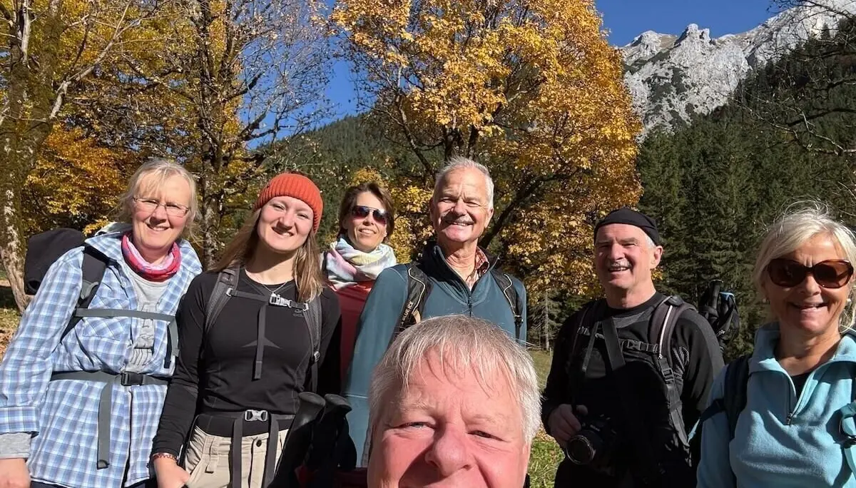 Sieben Erwachsene stehen vor herbstlich gefärbten Bäumen und Berggipfeln, lächeln in Wanderausrüstung in die Kamera. | © DAV Markt Schwaben · Foto: Markus Zimmermann