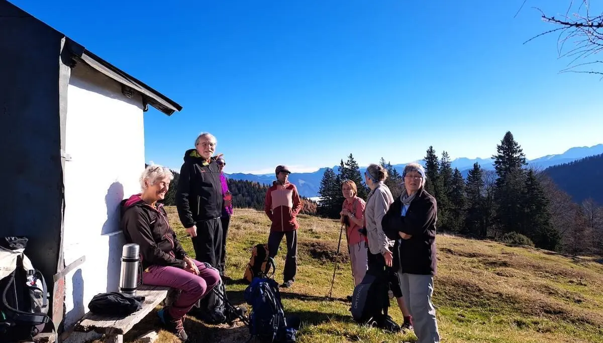Sieben Wandernde machen Rast vor einer kleinen weißen Hütte mit Bank. Einige stehen, eine Person sitzt, Rucksäcke und Thermosflasche liegen bereit. Im Hintergrund sind bewaldete Hügel und ferne Berggipfel unter klarem Himmel zu sehen – die Szene wirkt ruhig und gemeinschaftlich. | © DAV Markt Schwaben | Foto: Tanja Döhler