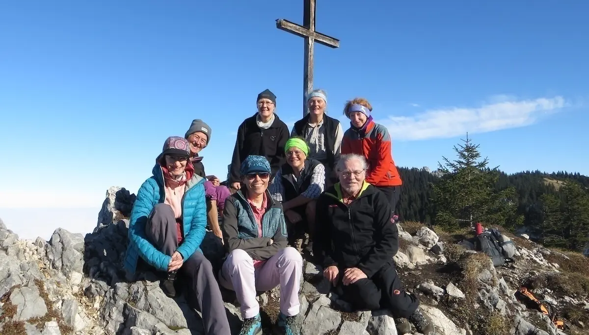 Acht Wandernde posieren auf einem felsigen Gipfel unter klarem Himmel vor einem großen Holzkreuz. Sie tragen Outdoor-Kleidung und wirken zufrieden nach dem Aufstieg. Im Hintergrund sind Nadelbäume und sanfte Hügel zu sehen – die Szene strahlt Gemeinschaft und Naturverbundenheit aus. | © DAV Markt Schwaben | Foto: Tanja Döhler