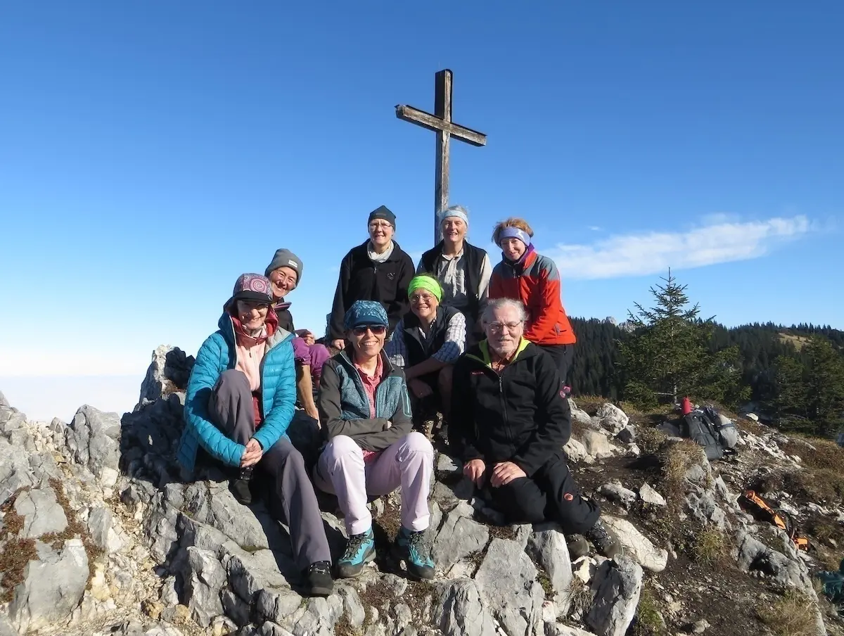 Acht Wandernde posieren auf einem felsigen Gipfel unter klarem Himmel vor einem großen Holzkreuz. Sie tragen Outdoor-Kleidung und wirken zufrieden nach dem Aufstieg. Im Hintergrund sind Nadelbäume und sanfte Hügel zu sehen – die Szene strahlt Gemeinschaft und Naturverbundenheit aus. | © DAV Markt Schwaben | Foto: Tanja Döhler