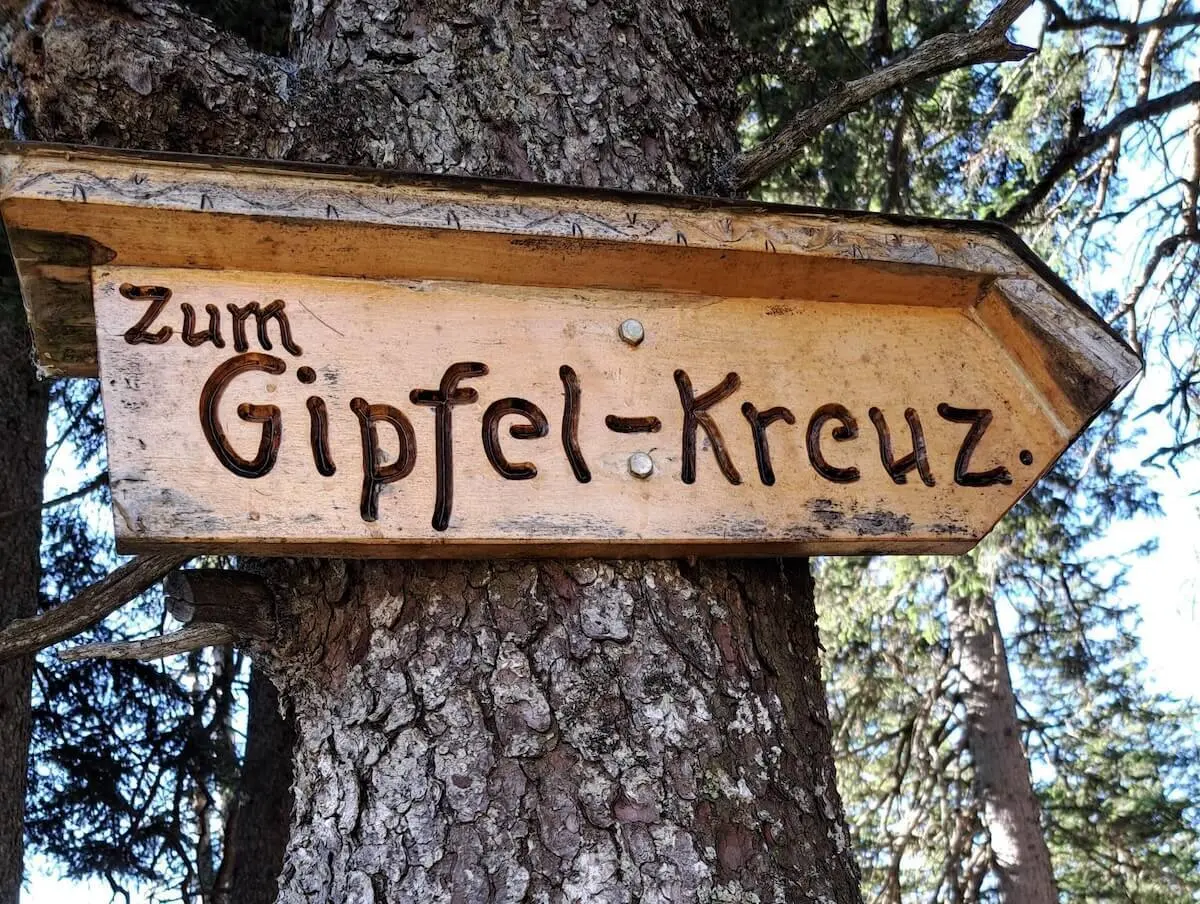 Ein hölzerner Richtungspfeil mit der Aufschrift „Zum Gipfel-Kreuz“ ist an einem Baum befestigt. Der Wegweiser zeigt nach rechts und fügt sich rustikal in die bewaldete Umgebung ein. Er markiert den Pfad zum Gipfel und vermittelt Orientierung und Vorfreude auf das Ziel. | © DAV Markt Schwaben | Foto: Tanja Döhler