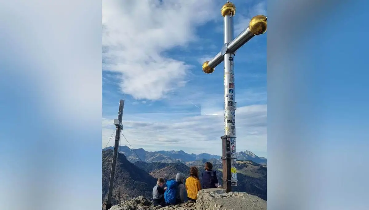 Vier Personen sitzen auf dem Gipfel des Kranzhorns vor einem großen Gipfelkreuz und blicken auf die umliegenden Berge. Das Bild zeigt Gemeinschaft und Naturerlebnis bei einer Familienwanderung. | © DAV Markt Schwaben | Foto: Maria Wenisch