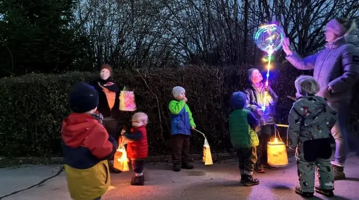 Mehrere Kinder und Erwachsene stehen draußen bei Dämmerung auf einem gepflasterten Platz. Die Kinder halten leuchtende Laternen, ein Erwachsener trägt einen bunten Leuchtstab. Alle sind warm angezogen, im Hintergrund sind Hecken und Bäume zu sehen. | © DAV Markt Schwaben · Foto: Lara Wolff