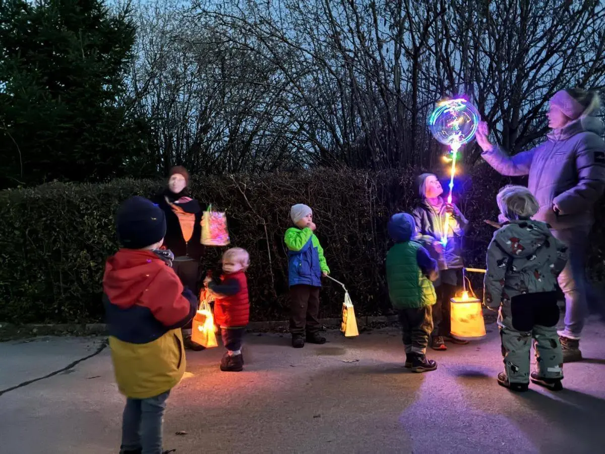 Mehrere Kinder und Erwachsene stehen draußen bei Dämmerung auf einem gepflasterten Platz. Die Kinder halten leuchtende Laternen, ein Erwachsener trägt einen bunten Leuchtstab. Alle sind warm angezogen, im Hintergrund sind Hecken und Bäume zu sehen. | © DAV Markt Schwaben · Foto: Lara Wolff