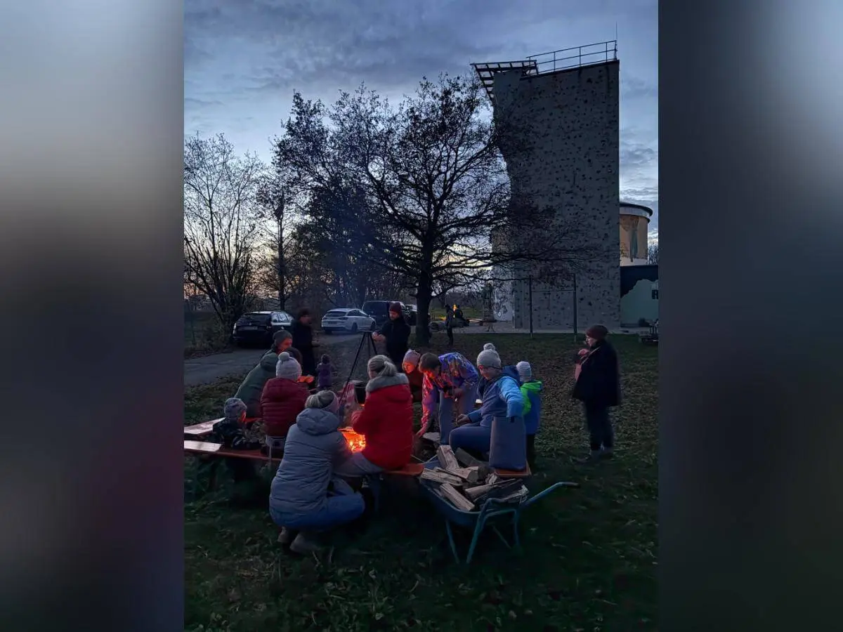 Mehrere Personen sitzen bei Dämmerung um ein Lagerfeuer, warm gekleidet mit Jacken und Mützen. Sie unterhalten sich oder schauen ins Feuer. Im Hintergrund stehen ein Kletterturm, ein rundes Gebäude und geparkte Autos. Die Bäume sind kahl, es wirkt herbstlich. | © DAV Markt Schwaben · Foto: Lara Wolff