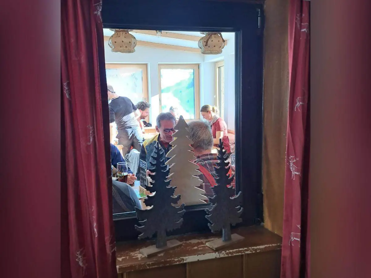 Blick durch Fenster mit roten Vorhängen auf hellen Gastraum, Personen sitzen und unterhalten sich, Deko im Vordergrund. | © DAV Markt Schwaben · Foto Tanja Döhler