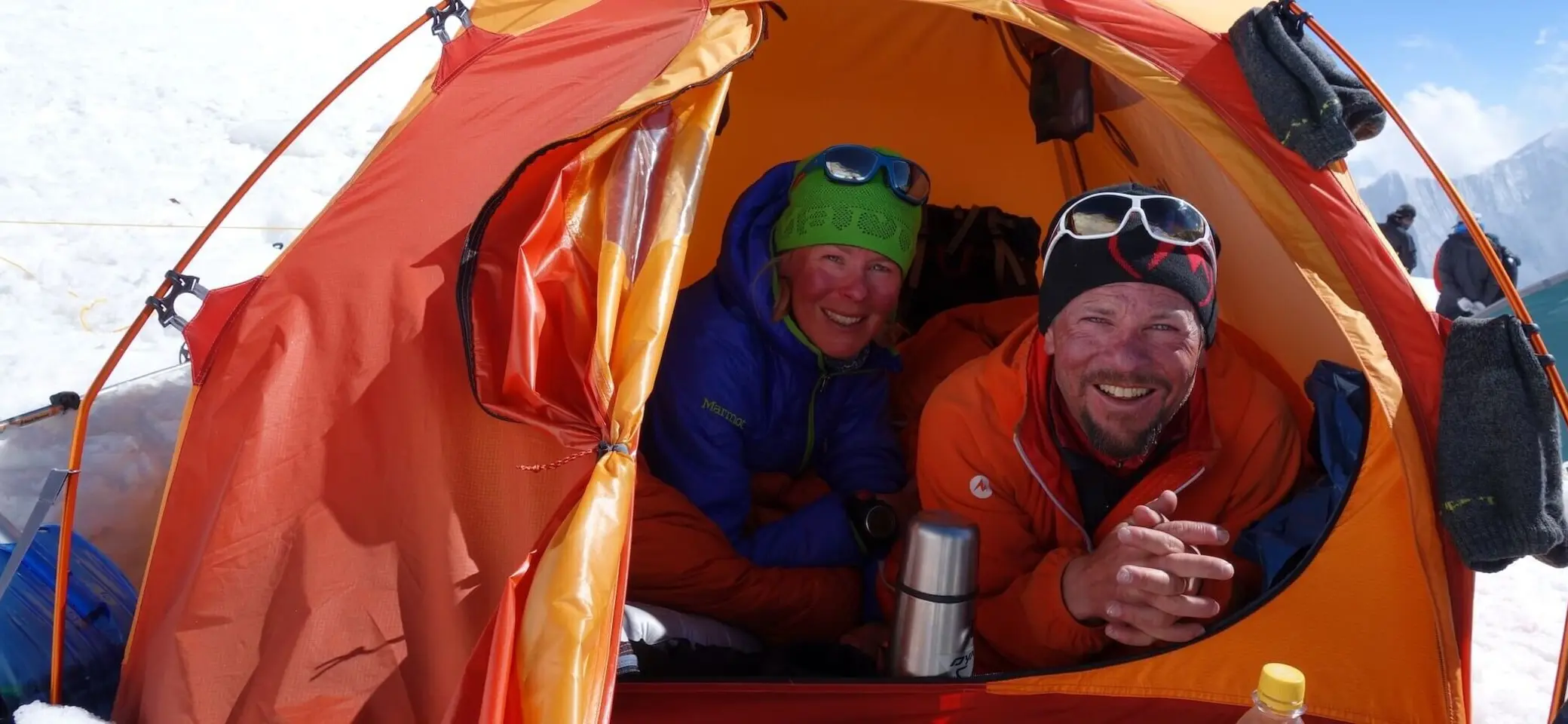 Zwei Personen sitzen in einem orangefarbenen Zelt auf einem verschneiten Berg, tragen Winterausrüstung und Sonnenbrillen. | © Alix von Melle