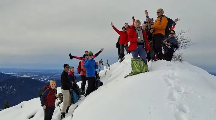 Eine Gruppe von Winterwandernden steht auf einem verschneiten Gipfel und posiert mit erhobenen Armen. Im Hintergrund sind ferne Bergketten und ein wolkenverhangener Himmel zu sehen. | © DAV Markt Schwaben · Foto Alfred Quiel