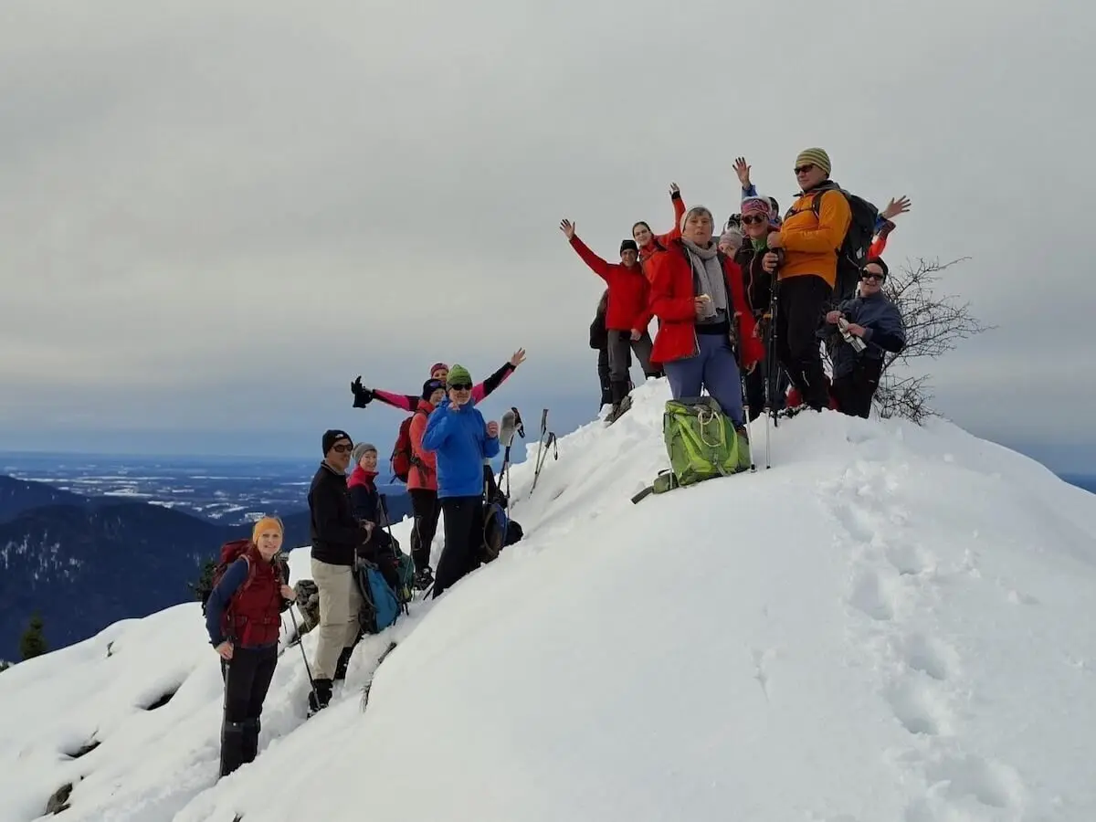 Eine Gruppe von Winterwandernden steht auf einem verschneiten Gipfel und posiert mit erhobenen Armen. Im Hintergrund sind ferne Bergketten und ein wolkenverhangener Himmel zu sehen. | © DAV Markt Schwaben · Foto Alfred Quiel