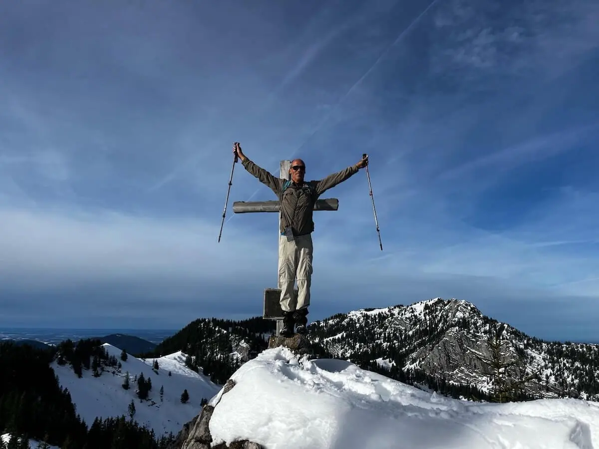 Ein Bergsteiger steht mit erhobenen Wanderstöcken neben einem Gipfelkreuz auf einem verschneiten Berg. Im Hintergrund erstreckt sich ein Panorama aus schneebedeckten Gipfeln unter blauem Himmel mit Wolken. | © DAV Markt Schwaben · Foto Markus Sellmeier