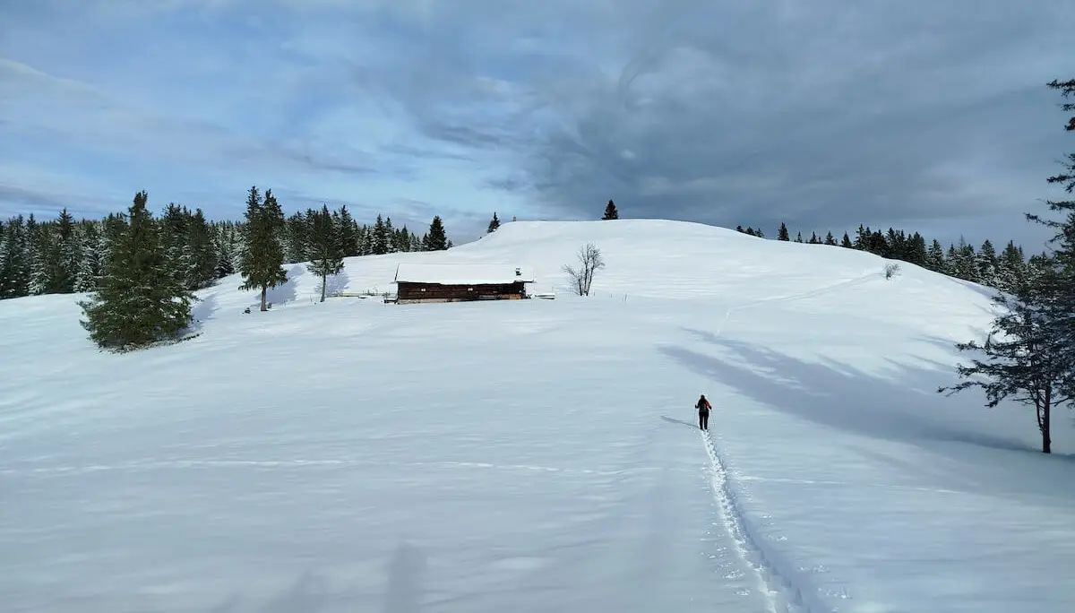 Eine Person wandert durch tiefen Schnee auf eine Holzhütte zu; umgeben von verschneiten Tannen und sanften Hügeln. | © DAV Markt Schwaben | Foto: Christine Strasser