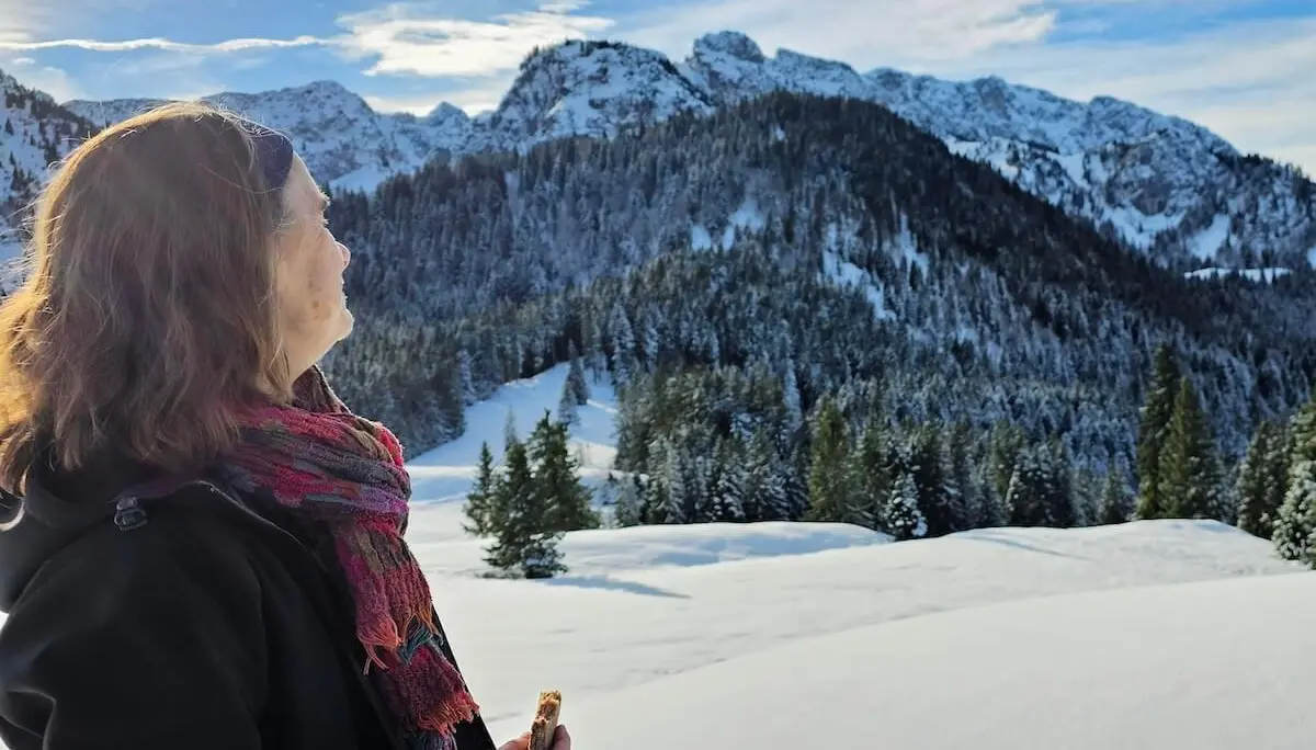 Eine Person steht im Schnee mit Müsliriegel in der Hand; im Hintergrund verschneite Berggipfel und Tannen im Sonnenlicht. | © DAV Markt Schwaben | Foto: Lutz Gründel
