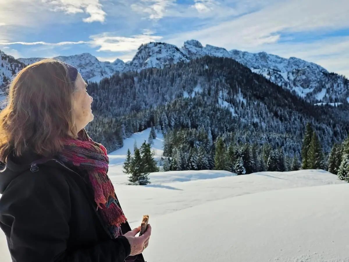Eine Person steht im Schnee mit Müsliriegel in der Hand; im Hintergrund verschneite Berggipfel und Tannen im Sonnenlicht. | © DAV Markt Schwaben | Foto: Lutz Gründel