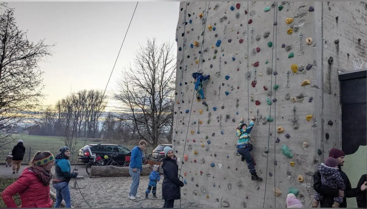 Zwei Kinder klettern gesichert an der bunten Kletterwand; Erwachsene stehen daneben, Bäume im Hintergrund. | © DAV Markt Schwaben · Familiengruppe