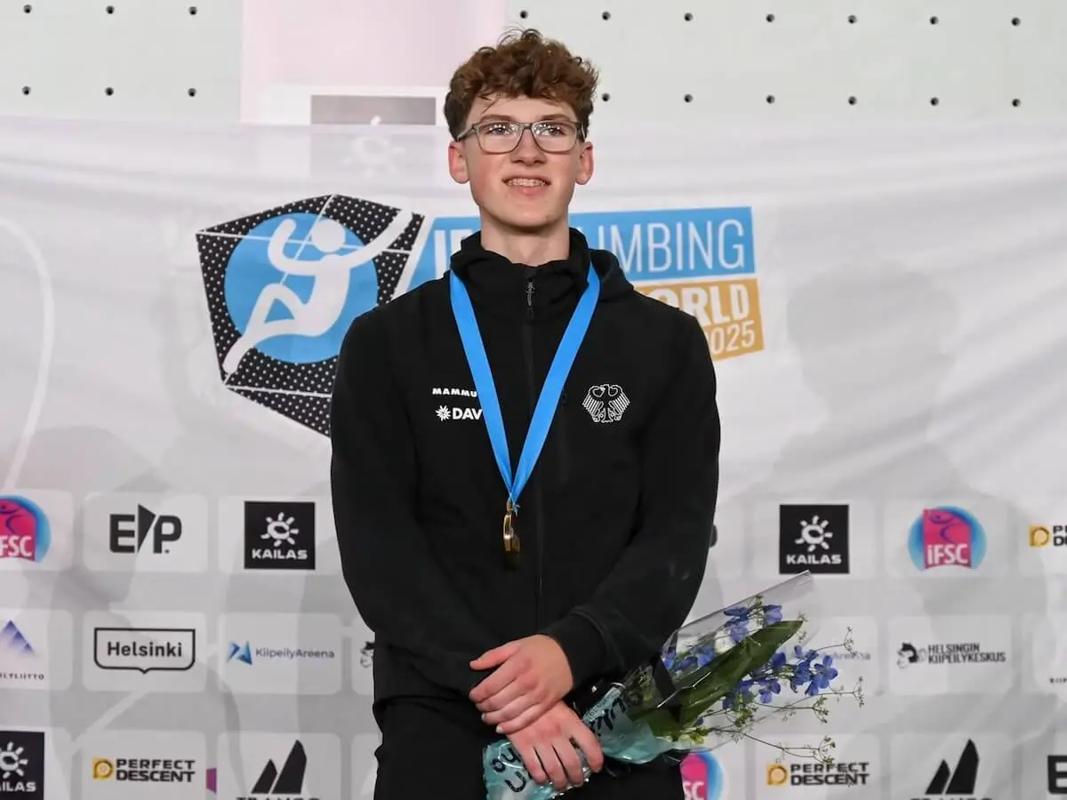 Aodhán Umlauf, 18, steht bei der U19‑WM‑Siegerehrung in Helsinki mit Goldmedaille und Blumenstrauß vor einer Sponsorwand. | © Foto: Richard Aspland / IFSC