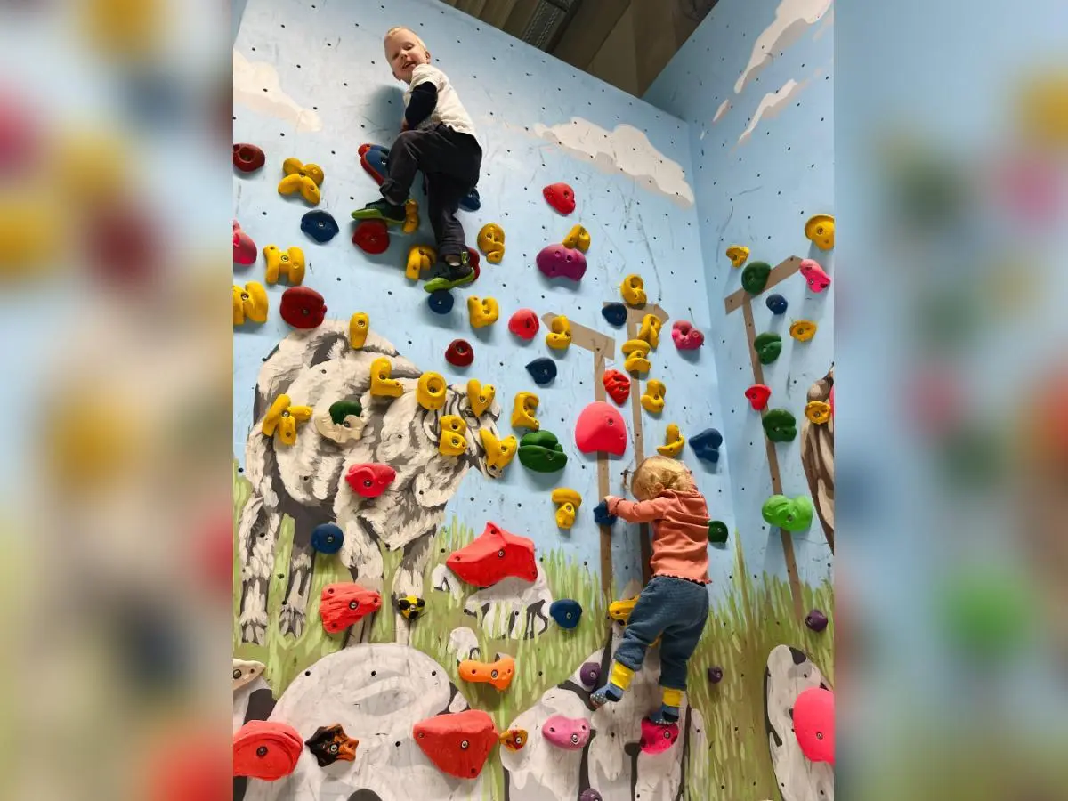 Zwei Kinder klettern an bunten Griffen vor einer Wand mit Tiermotiven; Schaf und Kuh im Hintergrund, Fokus auf Bewegung. | © DAV Markt Schwaben/Lara Wolff