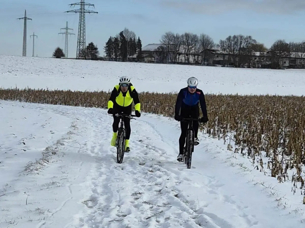 Zwei Radfahrer fahren nebeneinander auf verschneitem Feldweg; helle Kleidung hebt sich vom winterlichen Hintergrund ab. | © DAV Markt Schwaben/Miron Thoms
