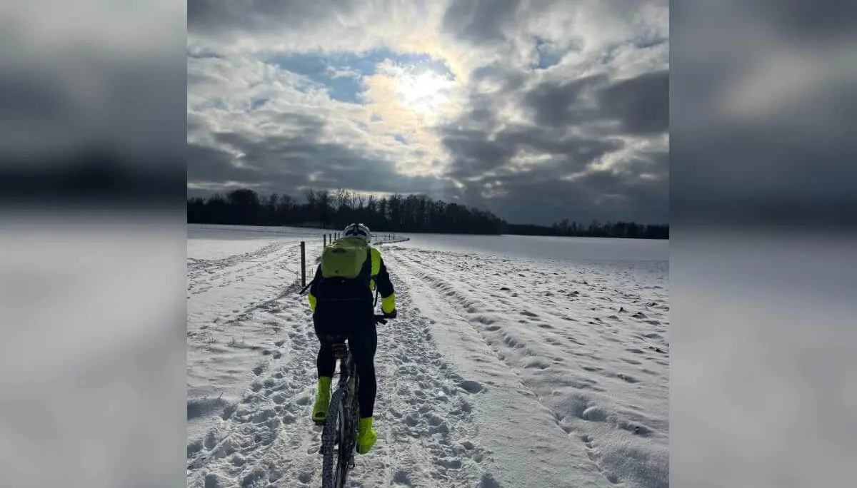 Eine Person fährt mit Mountainbike und gelber Jacke auf verschneitem Feldweg; Bäume und Sonne im wolkigen Hintergrund. | © DAV Markt Schwaben/Miron Thoms