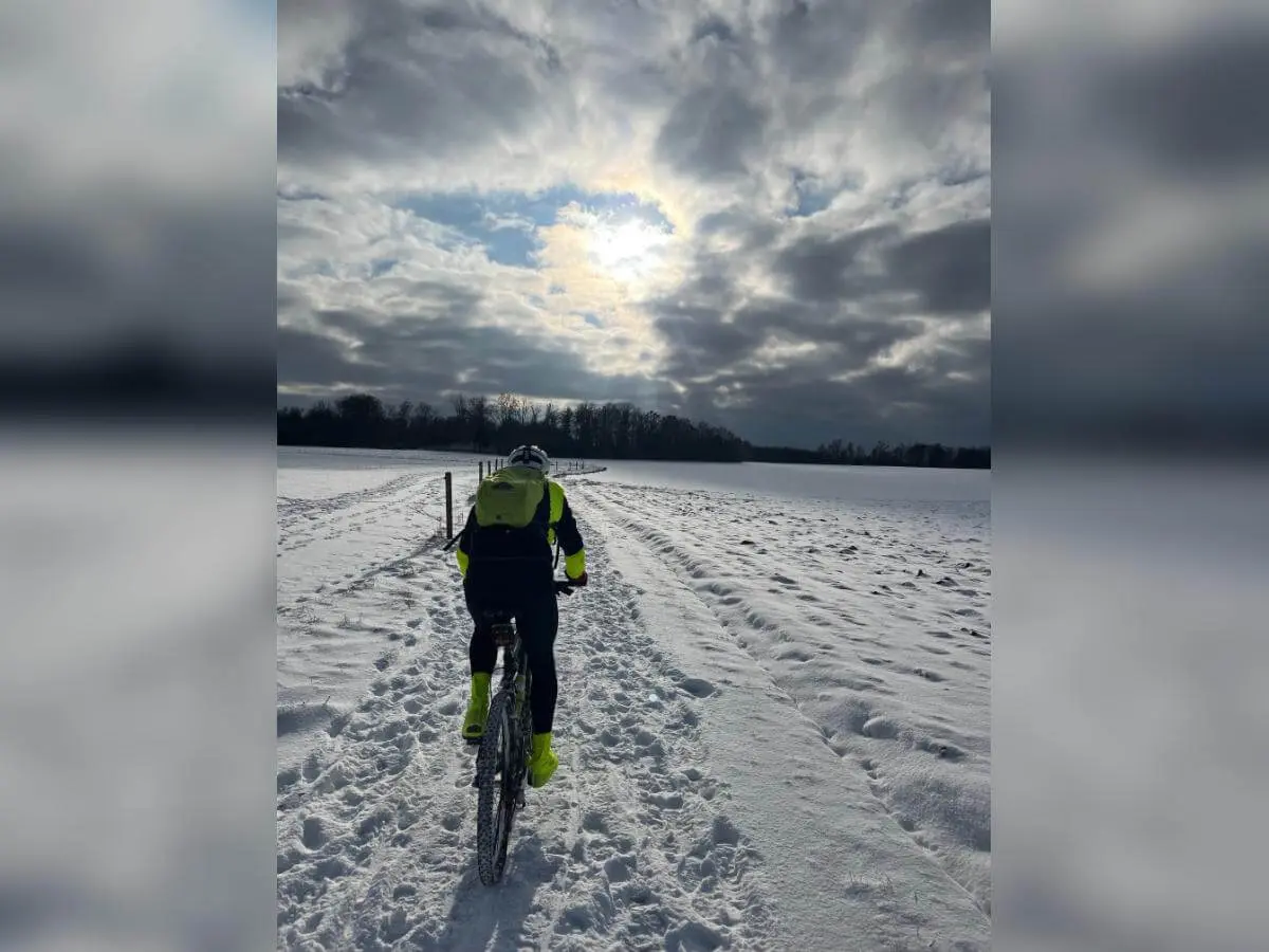 Eine Person fährt mit Mountainbike und gelber Jacke auf verschneitem Feldweg; Bäume und Sonne im wolkigen Hintergrund. | © DAV Markt Schwaben/Miron Thoms