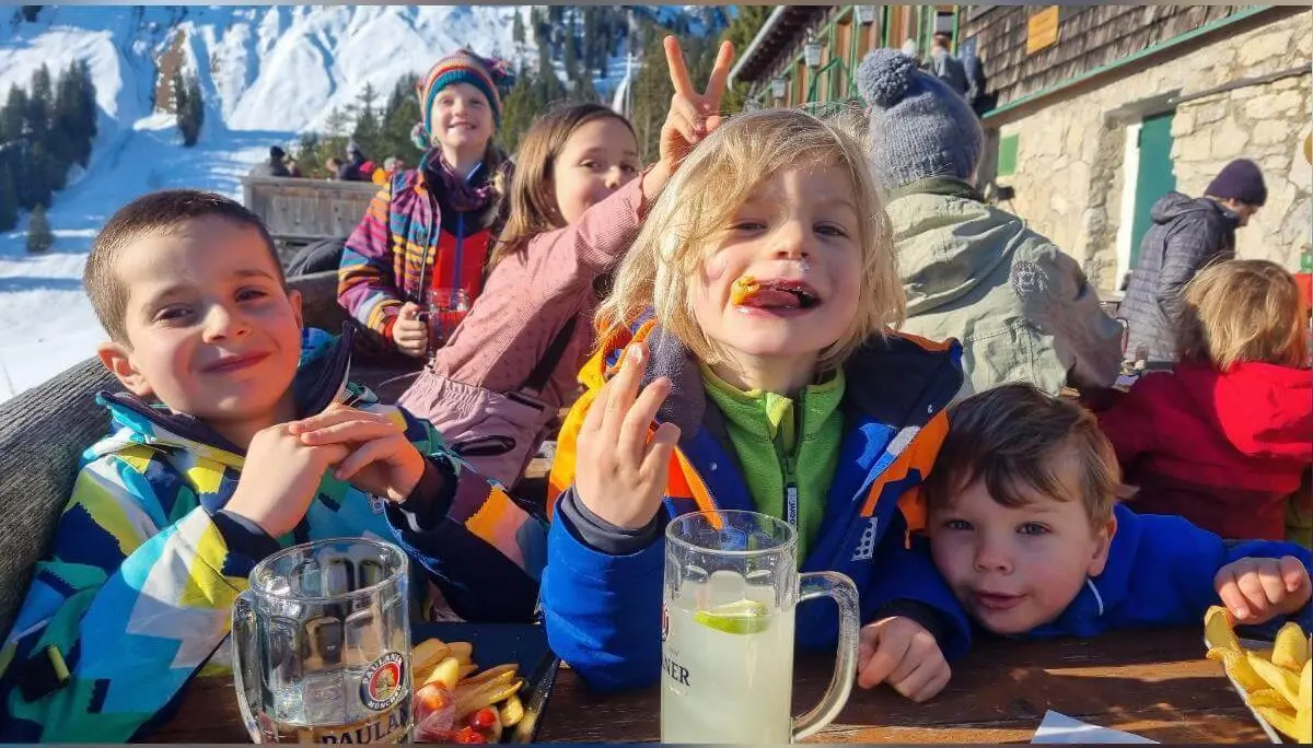 Kinder in bunter Winterkleidung essen draußen an einem Holztisch vor verschneiter Berghütte mit grünen Fensterläden. | © DAV Markt Schwaben/Silke Titze