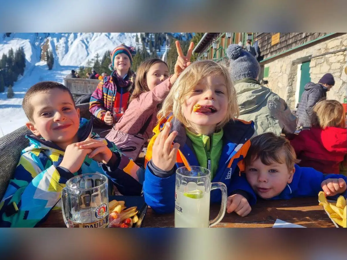 Kinder in bunter Winterkleidung essen draußen an einem Holztisch vor verschneiter Berghütte mit grünen Fensterläden. | © DAV Markt Schwaben/Silke Titze