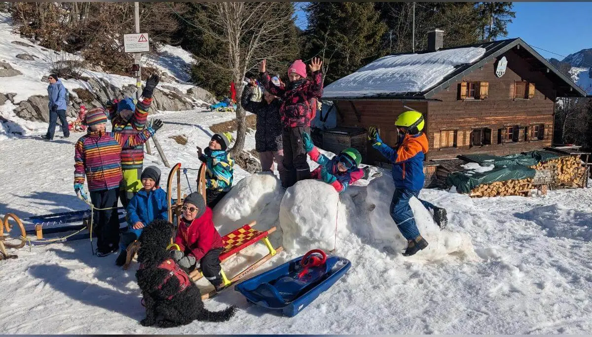 Kinder mit Helmen und Schlitten spielen im Schnee vor einer Berghütte; ein Hund läuft zwischen ihnen umher. | © DAV Markt Schwaben/Silke Titze