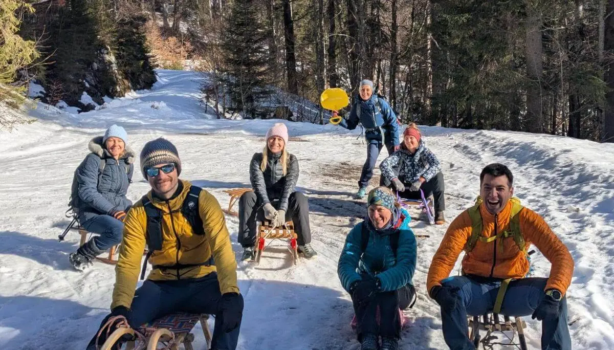 Acht Erwachsene sitzen mit Schlitten auf einem sonnigen Waldweg, umgeben von Schnee und Tannenbäumen. | © DAV Markt Schwaben/Silke Titze