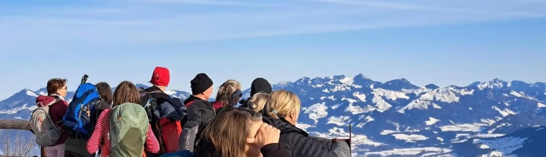 Gruppe Erwachsener steht im verschneiten Vordergrund an einem Holzgeländer und blickt über das weite Alpenpanorama im klaren Winterlicht. | © DAV Markt Schwaben/Erwin Matzinger