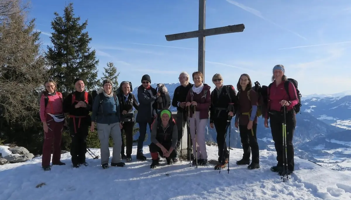 Elf Erwachsene stehen auf dem verschneiten Gipfel neben einem großen Holzkreuz und blicken vor klarer Bergkulisse in die winterlichen Alpen. | © DAV Markt Schwaben/Erwin Matzinger
