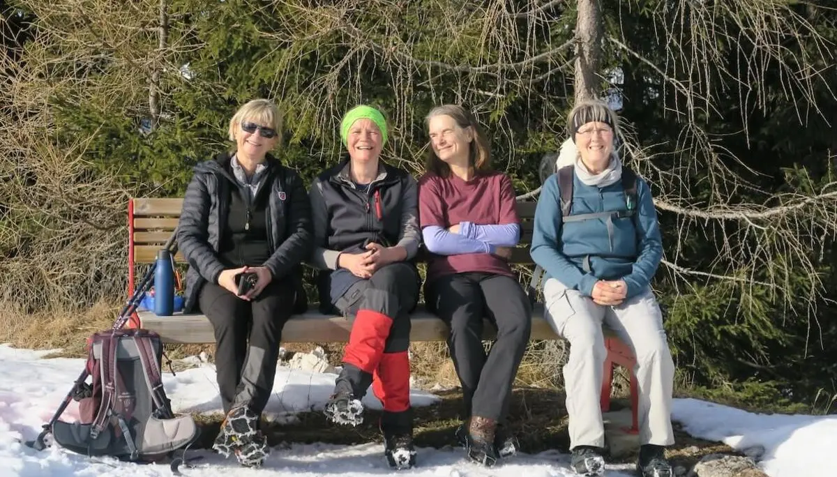 Vier Erwachsene sitzen in Winterkleidung auf einer Holzbank im Schnee, lachen entspannt und machen Rast zwischen verschneiten Bäumen. | © DAV Markt Schwaben/Erwin Matzinger