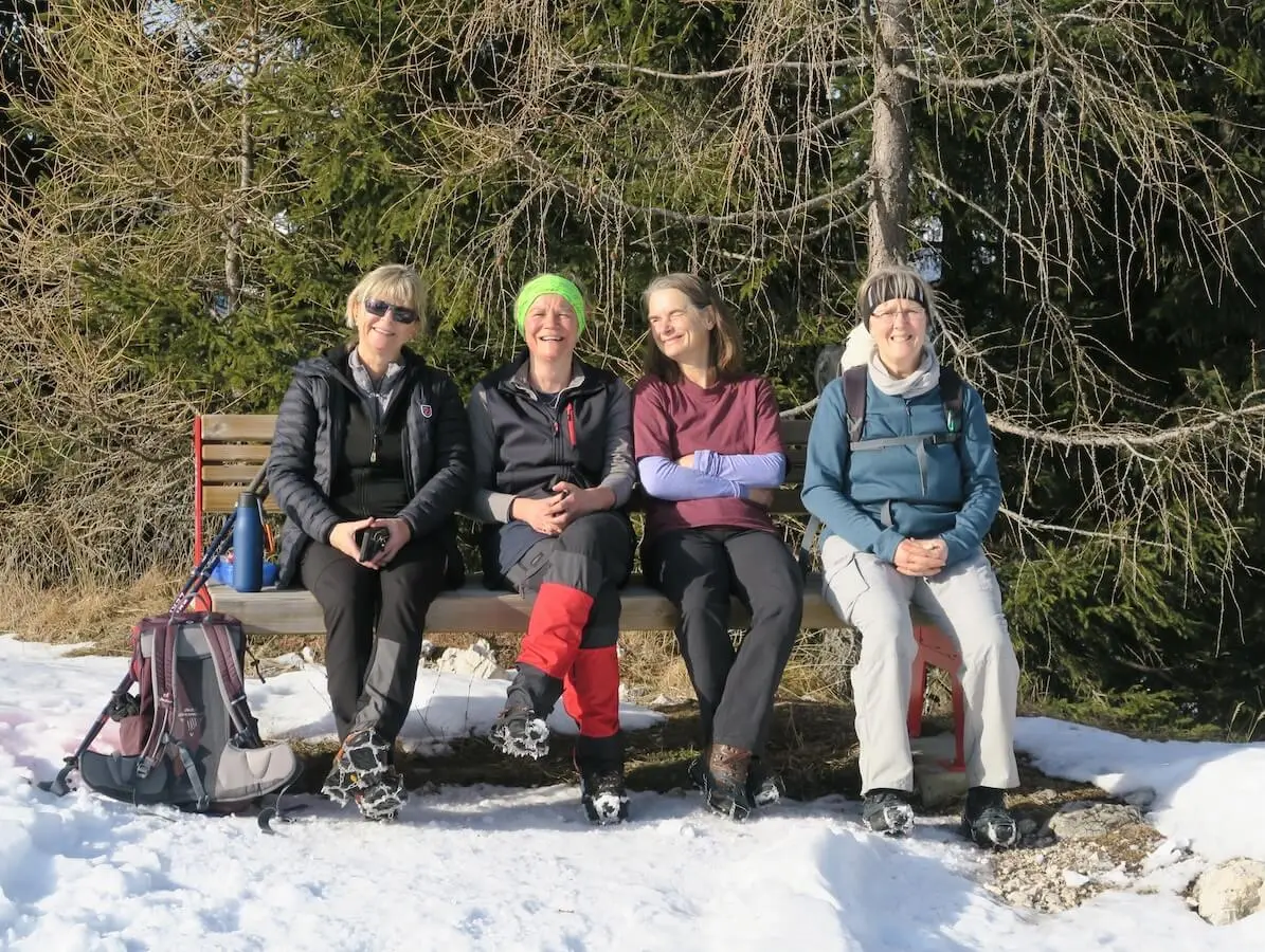 Vier Erwachsene sitzen in Winterkleidung auf einer Holzbank im Schnee, lachen entspannt und machen Rast zwischen verschneiten Bäumen. | © DAV Markt Schwaben/Erwin Matzinger