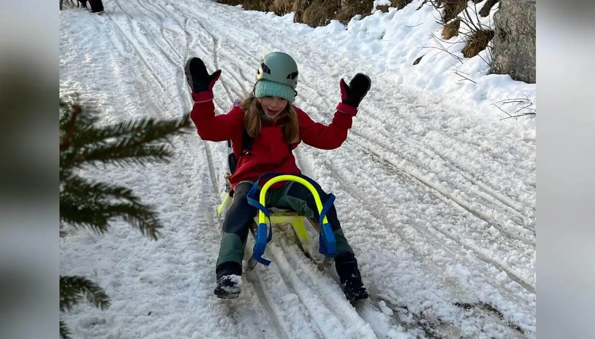 Ein Kind der Jugendgruppen Bergdohlen sitzt auf einem Schlitten auf einem verschneiten Waldweg, hebt jubelnd die Arme und trägt Winterkleidung sowie Helm. | © DAV Markt Schwaben / Wiebke Rauschenbach