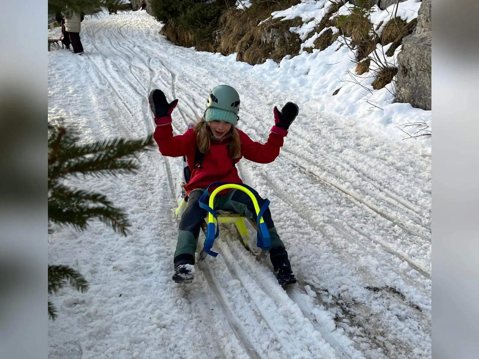 Ein Kind der Jugendgruppen Bergdohlen sitzt auf einem Schlitten auf einem verschneiten Waldweg, hebt jubelnd die Arme und trägt Winterkleidung sowie Helm. | © DAV Markt Schwaben / Wiebke Rauschenbach