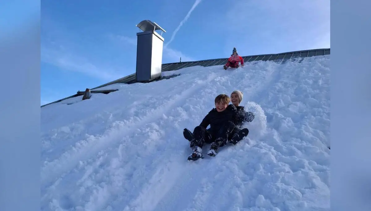 Zwei Jugendliche der Jugendgruppen Bergdohlen rodeln gemeinsam einen steilen, verschneiten Hang hinunter, im Hintergrund weitere Kinder und ein Gebäudedach unter blauem Himmel. | © DAV Markt Schwaben / Wiebke Rauschenbach