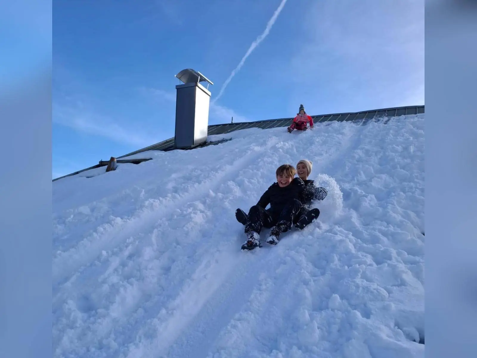 Zwei Jugendliche der Jugendgruppen Bergdohlen rodeln gemeinsam einen steilen, verschneiten Hang hinunter, im Hintergrund weitere Kinder und ein Gebäudedach unter blauem Himmel. | © DAV Markt Schwaben / Wiebke Rauschenbach