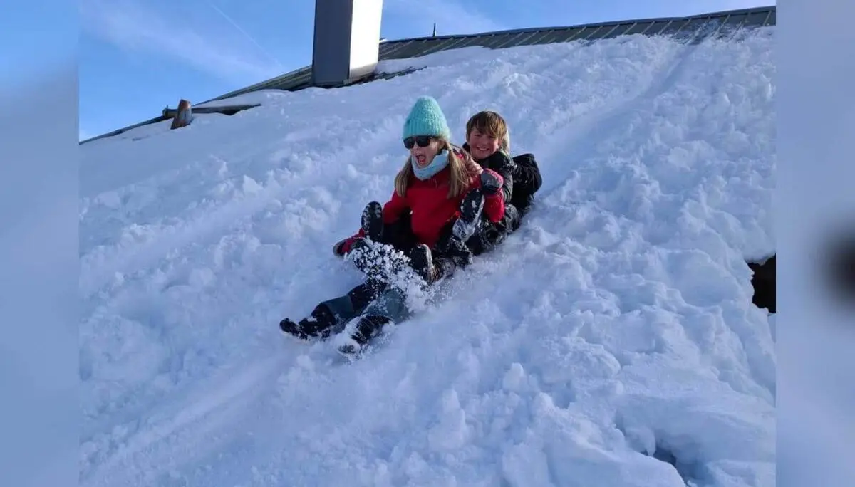 Zwei Jugendliche der Jugendgruppen Bergdohlen rodeln lachend gemeinsam einen verschneiten Hang hinunter, im Hintergrund Dach und klarer Himmel. | © DAV Markt Schwaben / Wiebke Rauschenbach