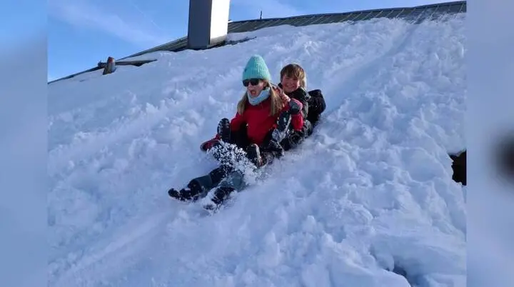 Zwei Jugendliche der Jugendgruppen Bergdohlen rodeln lachend gemeinsam einen verschneiten Hang hinunter, im Hintergrund Dach und klarer Himmel. | © DAV Markt Schwaben / Wiebke Rauschenbach