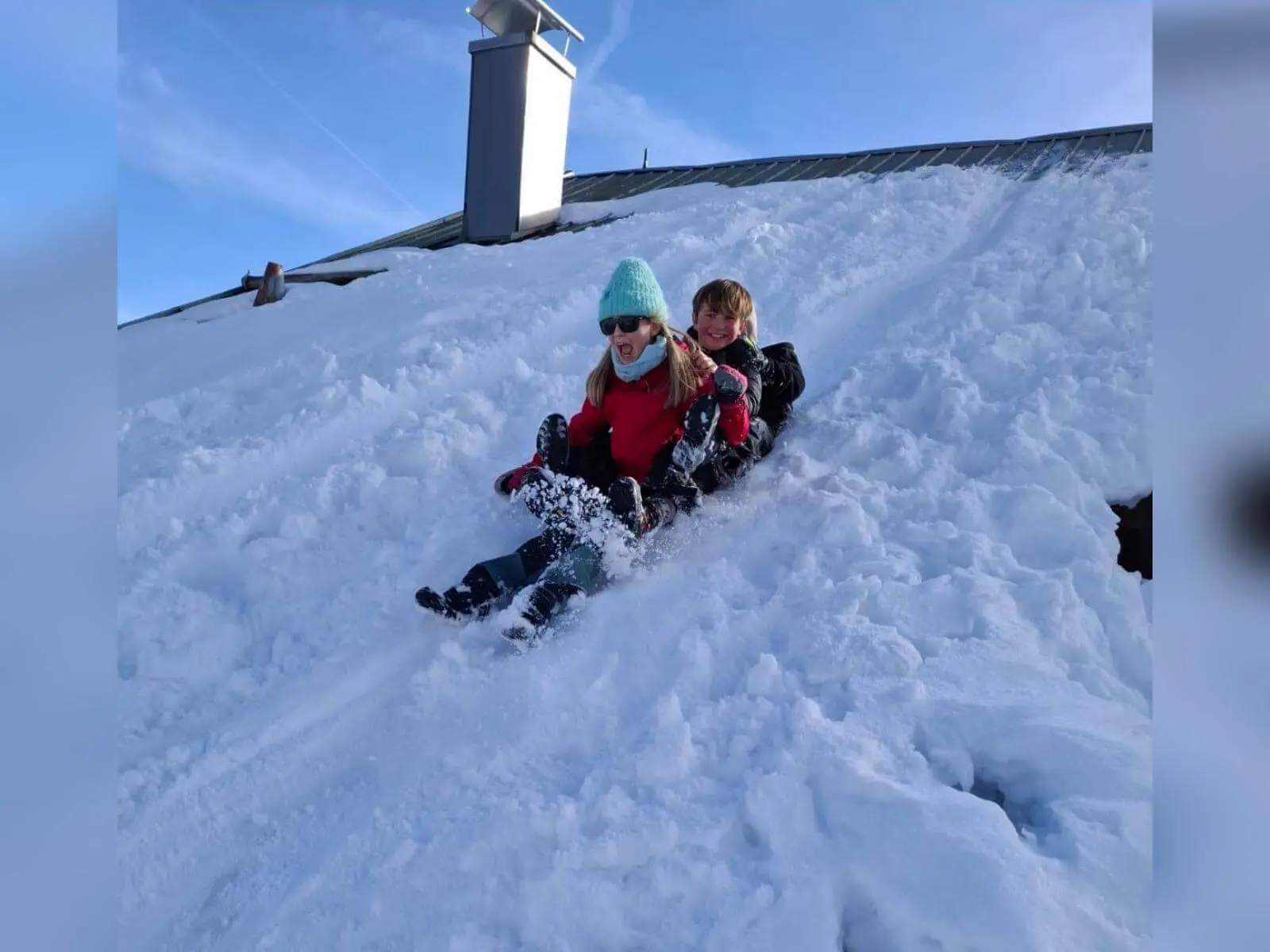 Zwei Jugendliche der Jugendgruppen Bergdohlen rodeln lachend gemeinsam einen verschneiten Hang hinunter, im Hintergrund Dach und klarer Himmel. | © DAV Markt Schwaben / Wiebke Rauschenbach