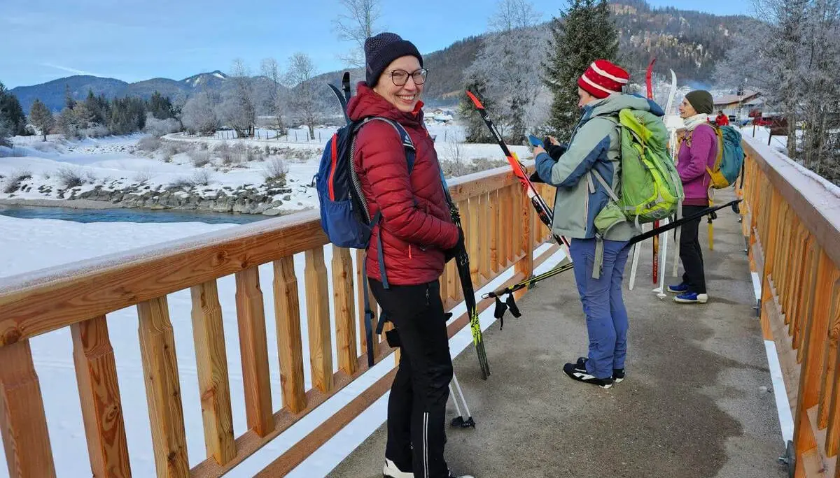 Mehrere Langläufer mit Skiern und Rucksäcken stehen auf einer Brücke über die verschneite Isar. Im Hintergrund sind winterliche Berge und Bäume zu sehen. | © DAV Markt Schwaben/Lutz Gründel