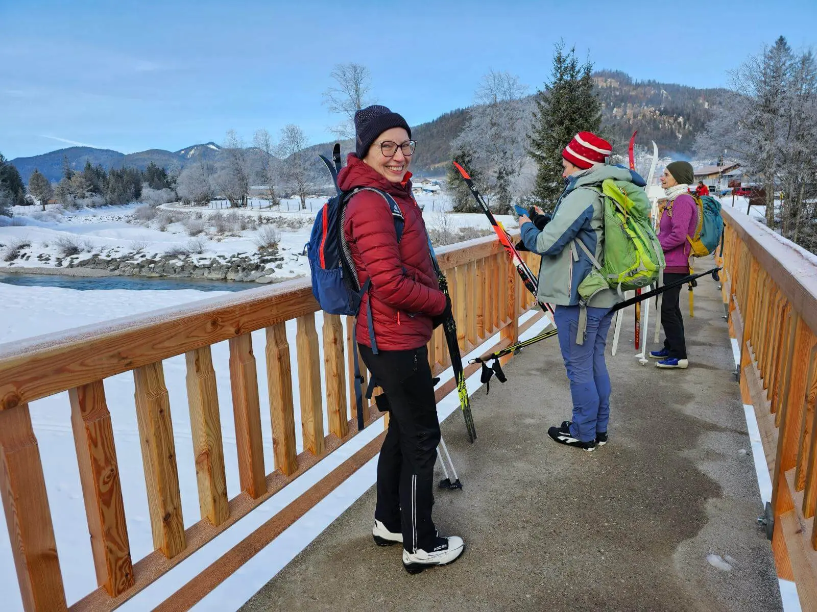 Mehrere Langläufer mit Skiern und Rucksäcken stehen auf einer Brücke über die verschneite Isar. Im Hintergrund sind winterliche Berge und Bäume zu sehen. | © DAV Markt Schwaben/Lutz Gründel