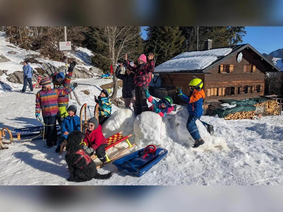 Kinder mit Helmen und Schlitten spielen im Schnee vor einer Berghütte; ein Hund läuft zwischen ihnen umher. | © DAV Markt Schwaben | Foto: Silke Titze