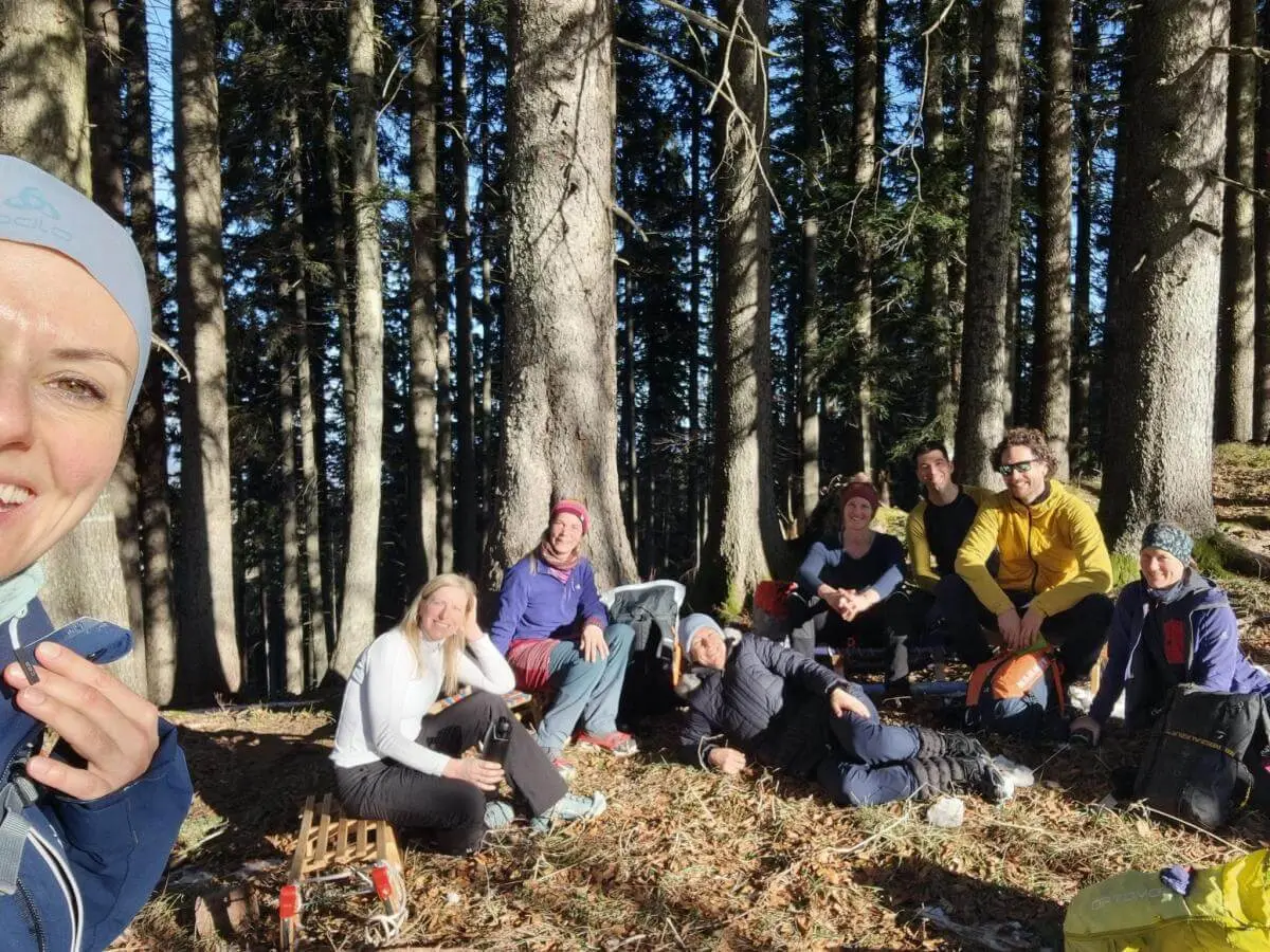 Acht Erwachsene sitzen entspannt im Wald zwischen Rucksäcken und Sonnenstrahlen, eine Person macht ein Selfie. | © DAV Markt Schwaben | Foto: Silke Titze