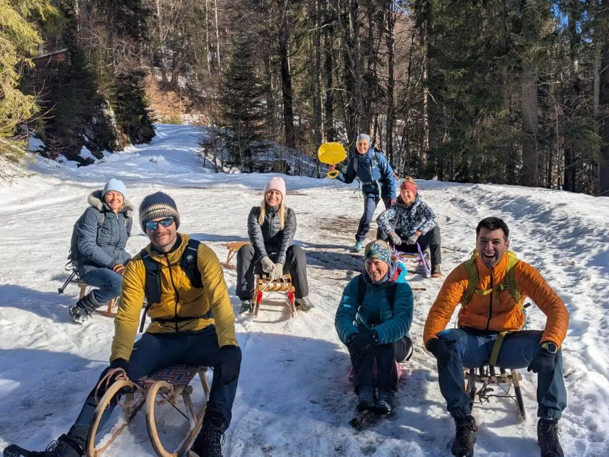 Acht Erwachsene sitzen mit Schlitten auf einem sonnigen Waldweg, umgeben von Schnee und Tannenbäumen. | © DAV Markt Schwaben | Foto: Silke Titze