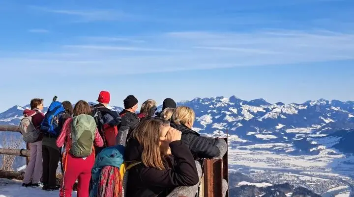 Gruppe Erwachsener steht im verschneiten Vordergrund an einem Holzgeländer und blickt über das weite Alpenpanorama im klaren Winterlicht. | © DAV Markt Schwaben / Erwin Matzinger