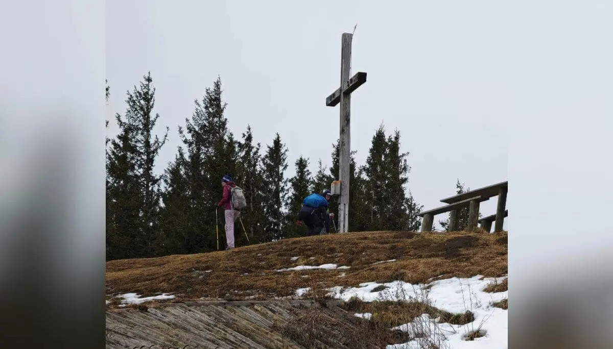 Zwei Wandernde stehen am hölzernen Gipfelkreuz auf einem teils verschneiten Hügel; einer kniet am Sockel, dahinter Tannen und bewölkter Himmel. | © DAV Markt Schwaben/Erwin Matzinger