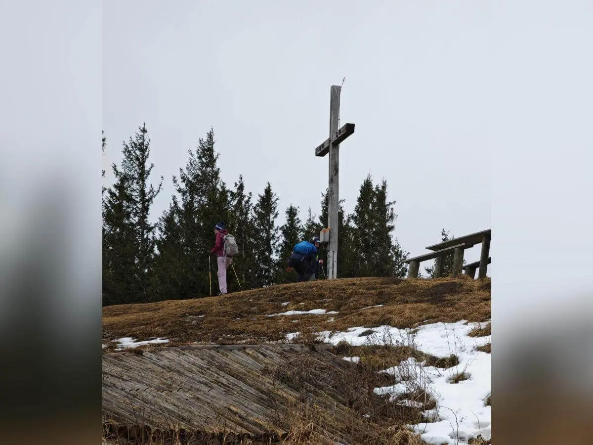 Zwei Wandernde stehen am hölzernen Gipfelkreuz auf einem teils verschneiten Hügel; einer kniet am Sockel, dahinter Tannen und bewölkter Himmel. | © DAV Markt Schwaben/Erwin Matzinger