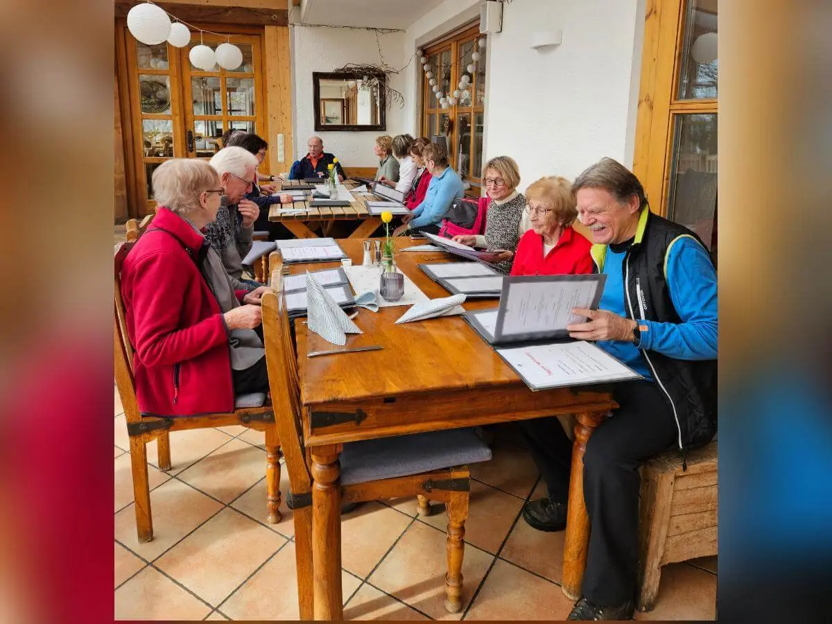 Seniorinnen und Senioren sitzen in einer warmen Stube an zwei Holztischen und lesen Speisekarten; durch große Fenster fällt Tageslicht. | © DAV Markt Schwaben/Hubert Inhofer