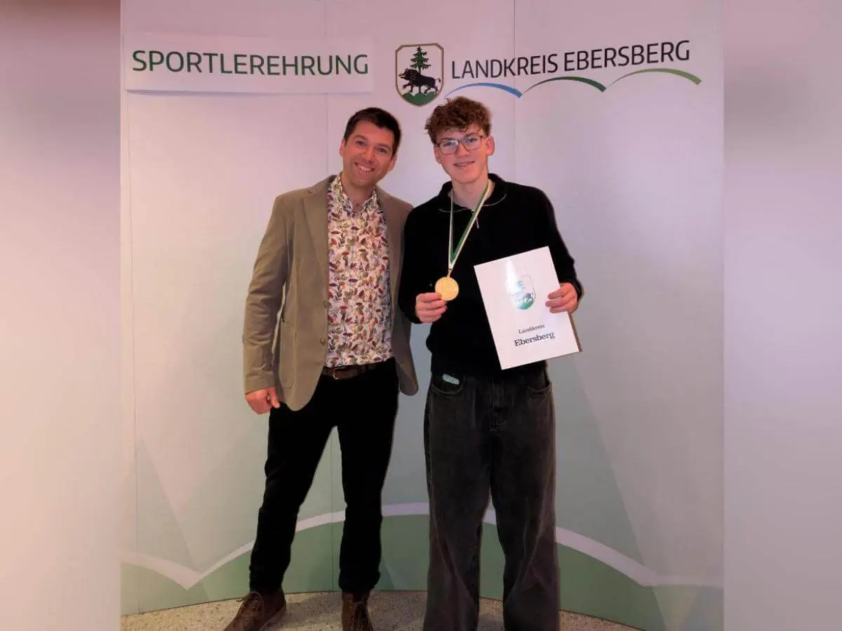 Zwei Personen stehen vor einem Banner mit der Aufschrift ‚Sportlerehrung Landkreis Ebersberg‘. Die geehrte Person trägt eine Goldmedaille um den Hals und hält eine Urkunde in den Händen.  | © Sportlerehrung Landkreis Ebersberg