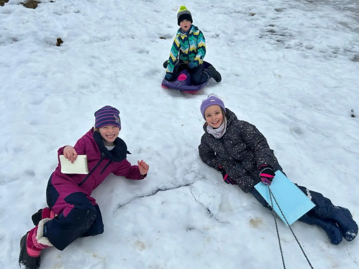 Drei Kinder spielen im Schnee; zwei liegen lachend vorn mit Notizbuch und Mappe, hinten sitzt ein Kind auf einem Schlitten am Fundort ihrer Entdeckung. | © DAV Markt Schwaben